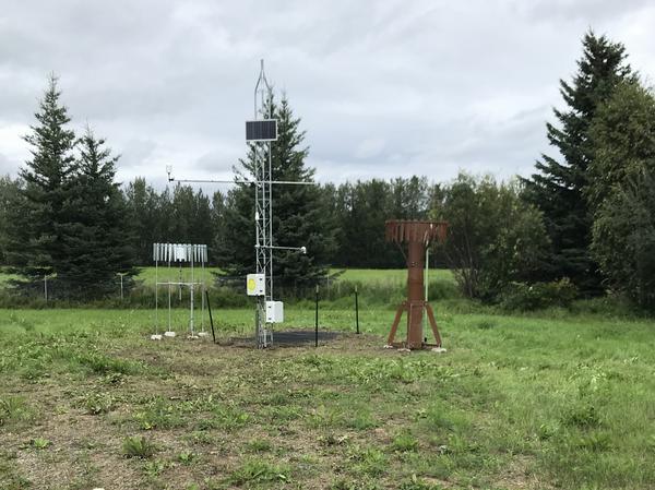 A tall metal scaffold holding science instruments and a solar panel stands between two circular instruments in a grassy field. Evergreen trees are in the background. 