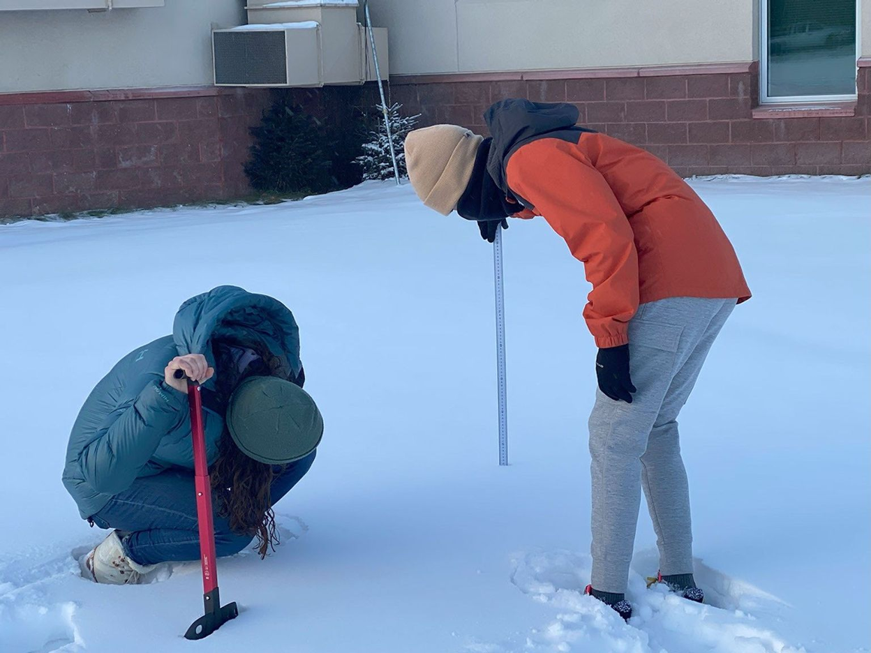 Two women in winter coats and hats are measuring snow depth outside of a brick building. One woman holds a tape measure in the snow, and the other is crouched down looking at the tape measure to record the depth of the snow. The crouching woman also holds a snow shovel. 