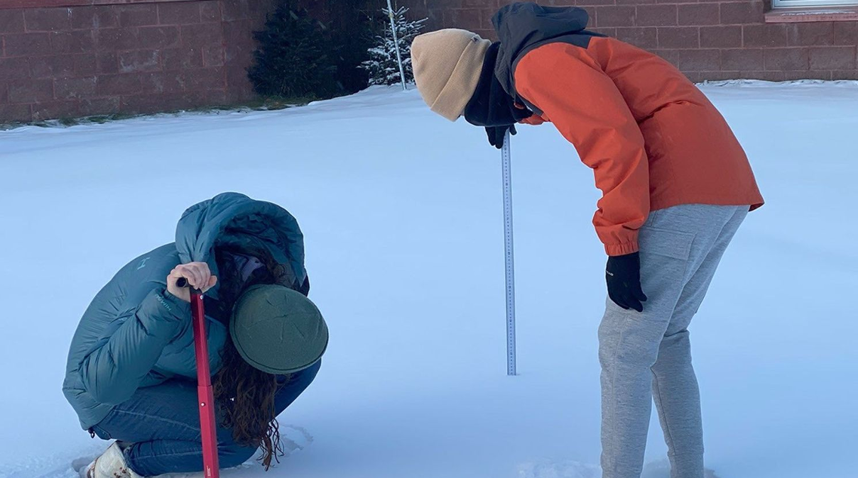 Two women in winter coats and hats are measuring snow depth outside of a brick building. One woman holds a tape measure in the snow, and the other is crouched down looking at the tape measure to record the depth of the snow. The crouching woman also holds a snow shovel.