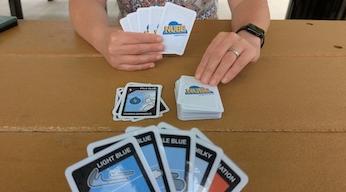 A person holds a hand of NUBE cards, about to draw a new card from the deck, which is next to the discard pile. In the foreground you can see the hand of the person they are playing with.