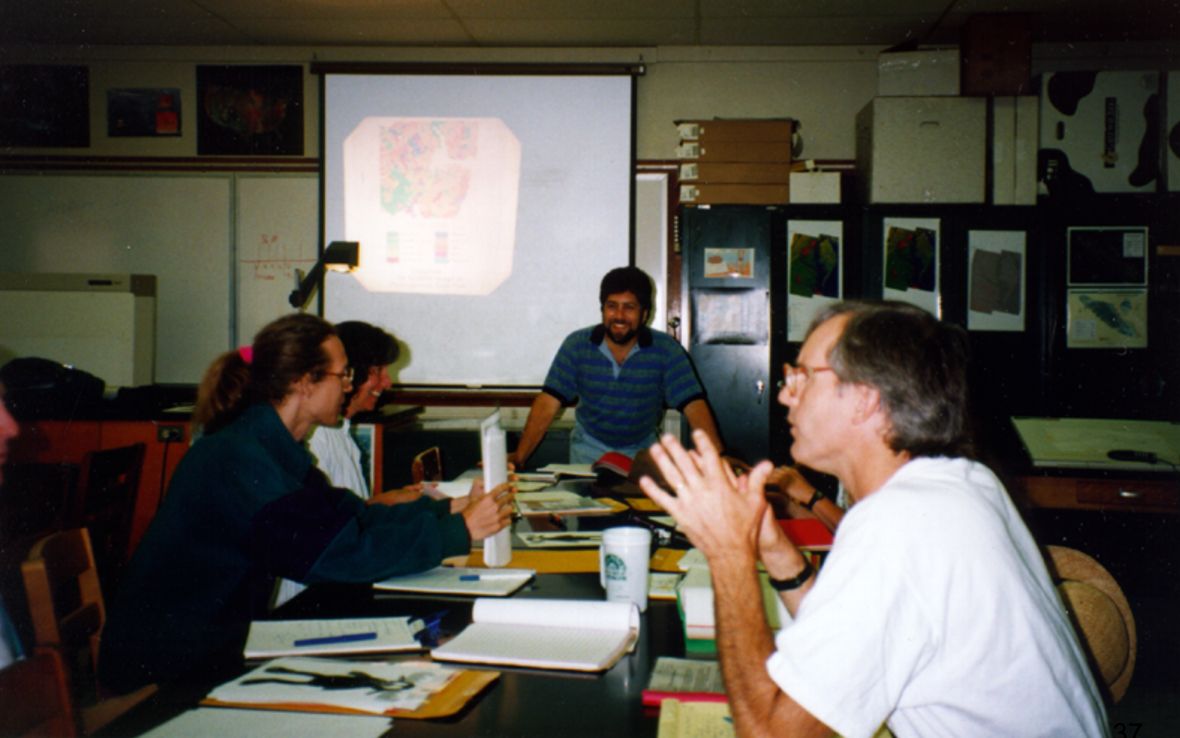A group of people sit around a long table looking toward a man standing at the top of the table. Behind him, an overhead projector casts a land cover classification image on a screen. 