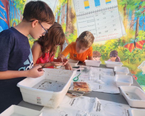 young campers gather around a table covered with tubs, nets, and identification keys to identify freshwater macroinvertebrates. The Earth Around Us water tent is in the background with colorful murals depicting a river ecosystem in three seasons.