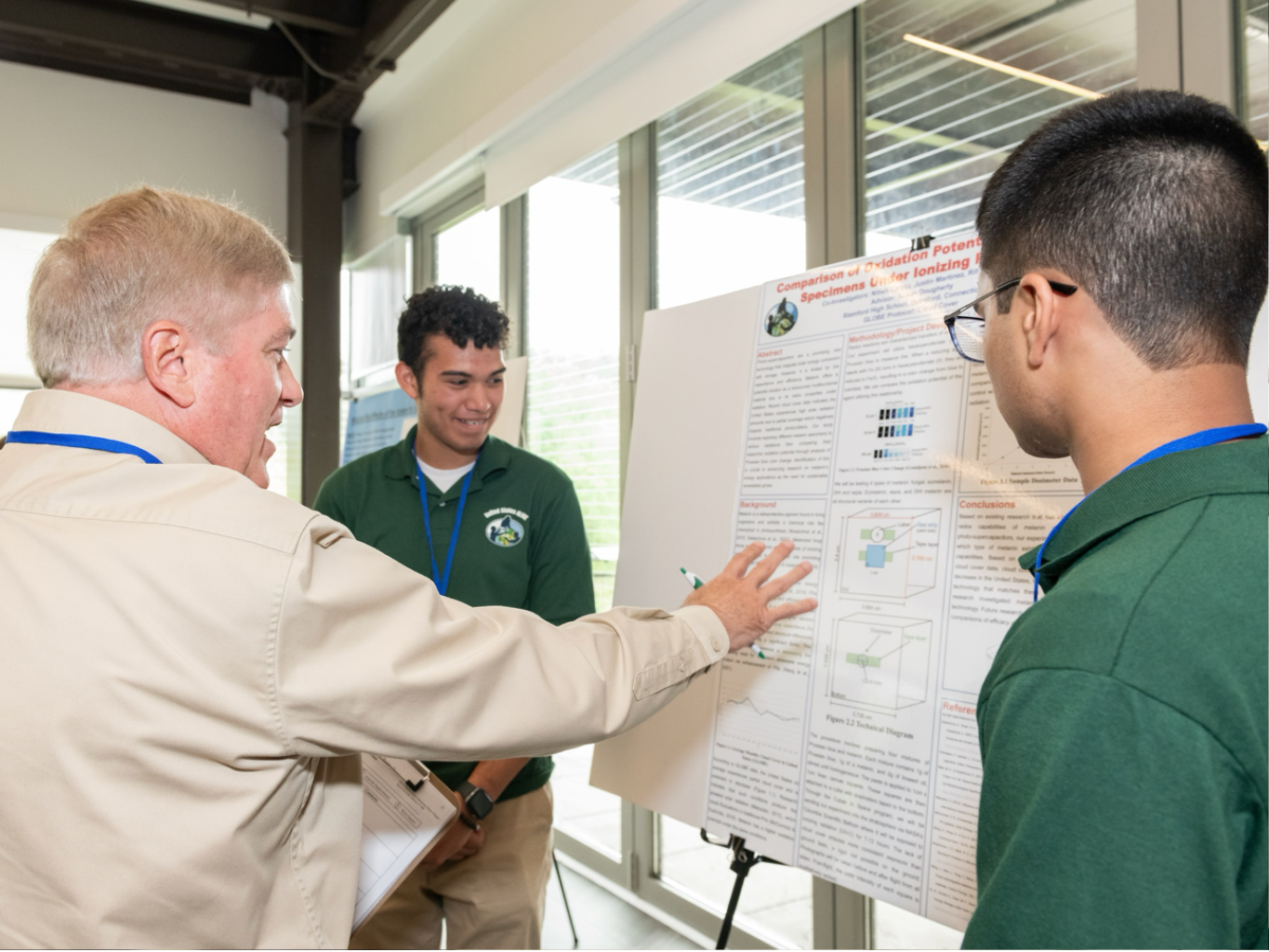 a STEM professional gestures at a research poster as he speaks with two high school-aged students about their GLOBE research